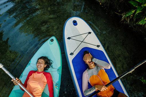 Photo of two teenage girls lying on their stand-up paddleboards and having a great time; teenagers hanging out in nature and enjoying their summer vacation.