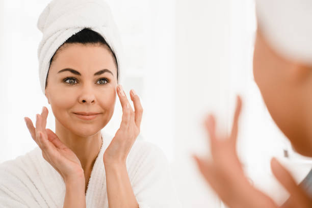 Good-looking caucasian middle-aged woman in turban and spa bathrobe after taking shower looking at the mirror while applying beauty creme moisturizer for anti-age anti-wrinkle effect stock photo