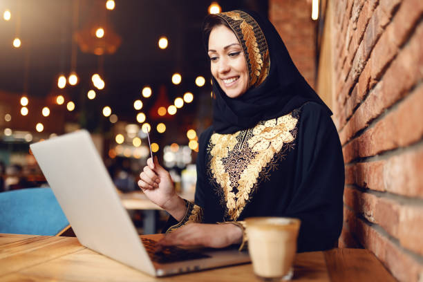Gorgeous muslim woman with toothy smile dressed in traditional wear using laptop for on-line shopping while sitting in cafeteria. On desk coffee. Gorgeous muslim woman with toothy smile dressed in traditional wear using laptop for on-line shopping while sitting in cafeteria. On desk coffee. hot middle eastern women stock pictures, royalty-free photos & images
