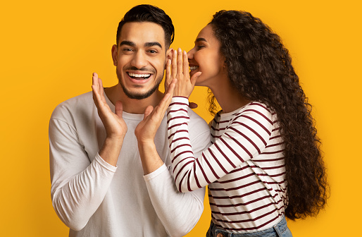 Gossips Concept. Lady Sharing Secret With Her Excited Arab Boyfriend, Joyful Middle Eastern Man Raising Hands In Amazement While Standing Together Over Yellow Studio Background, Empty Space