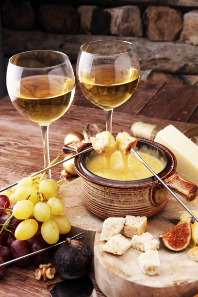 Gourmet Swiss fondue dinner on a winter evening with assorted cheeses on a board alongside a heated pot of cheese fondue with two forks dipping bread and white wine behind in a tavern or restaurant. stock photo