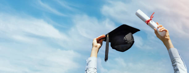graduate with diploma and cap in hands on blue sky background celebrating college graduation. banner copy space stock photo
