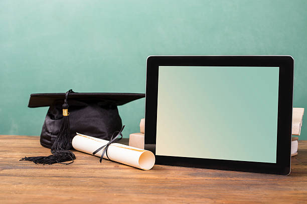 Graduation!  Cap, motarboard, diploma, digital tablet on desk.  Education. Spring graduation time.  A black cap/motarboard with black tassel on school desk beside a diploma, and digital tablet.   All objects lie on a rustic wooden desk with green chalkboard in background.  No people.  Education themes. online degree stock pictures, royalty-free photos & images