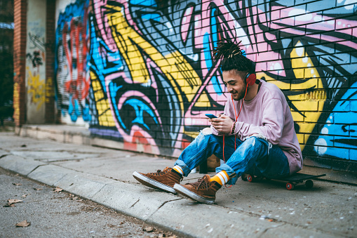 Young caucasian graffiti artist listens to music and using phone while sitting on a skateboard.