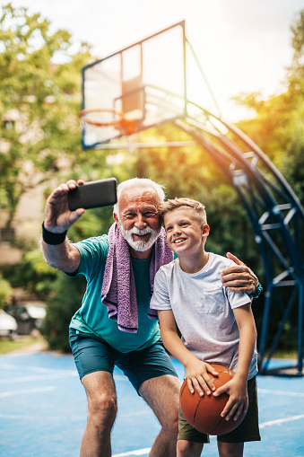 Grandfather and his grandson enjoying and playing together on basketball court. Grandfather uses smart phone to take selfie photo.