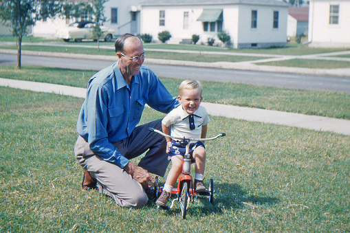 Grandfather helping grandson learn to ride tricycle. Iowa, USA 1953. Kodachrome scanned film with grain.