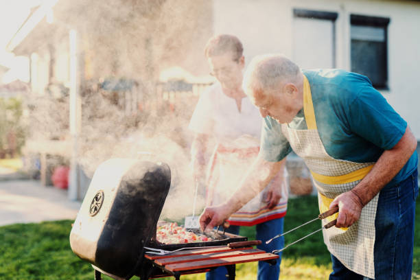 grandparents with aprons grilling meat and vegetables on sticks while standing in backyard at spring. - idosos aquecedor imagens e fotografias de stock