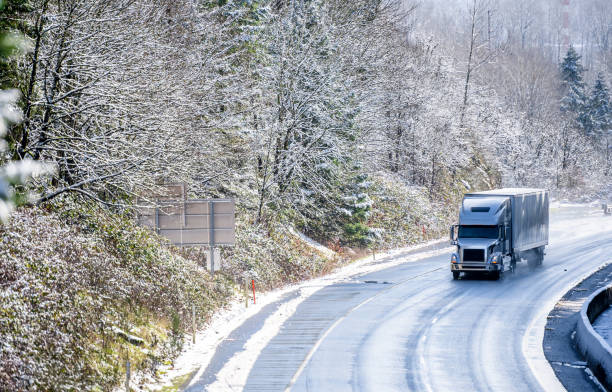 Gray big rig semi truck with covered semi trailer transporting cargo on winter snowing road with wet surface stock photo