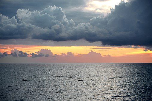 Gray-blue dramatic clouds above yellow sunlight and dark shine sea surface with kayaks