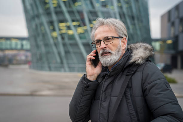 A gray-haired bearded businessman in a green winter jacket with glasses is phoning his cell phone while walking next to a glass office building. stock photo