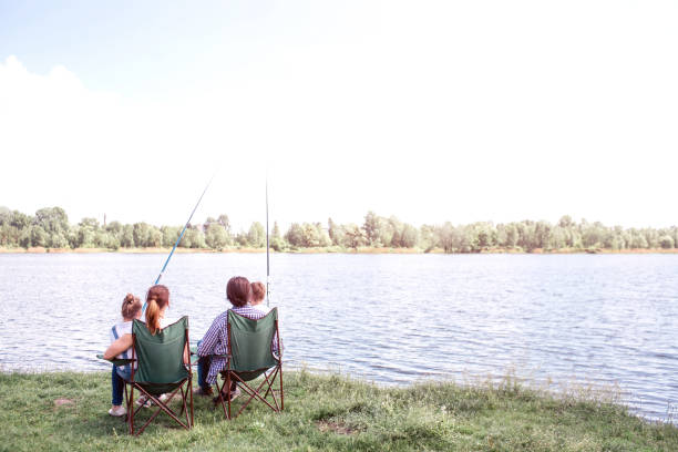 Great view of big river flowing. There is a family sitting by its shore and enjoying the moment. They are holding kids on the lap. Children are holding fish rods in hands. stock photo