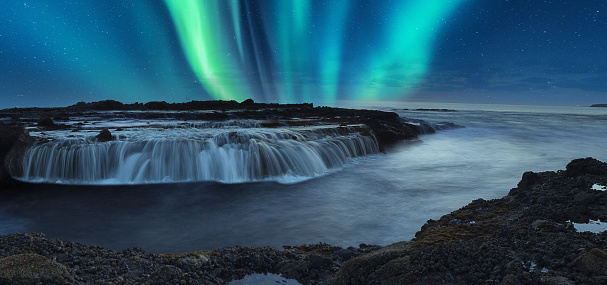 Green Aurora borealis shimmers over the ocean water as it cascades over rocks in Reykjavik, Iceland.
