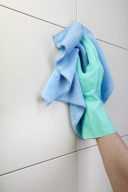 Green-gloved hand wiping shower tiles with blue cloth stock photo