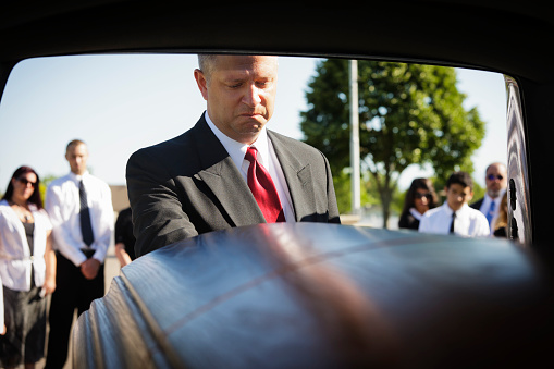 A grieving man standing at the rear of a hearse looking down at the casket.  Taken from within the hearse.