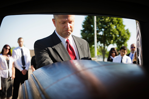 A grieving man standing at the rear of a hearse looking down at the casket. Taken from within the hearse.