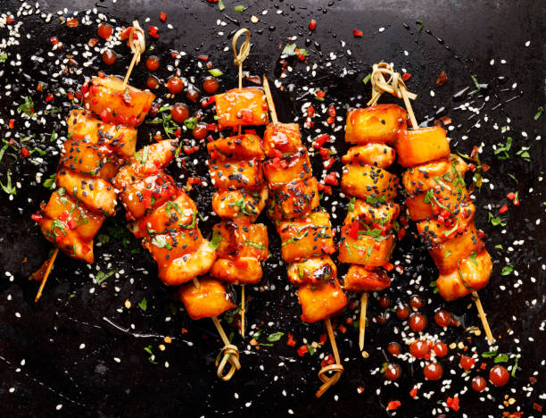 Grilled skewers with pineapple fruit and chicken meat  with sriracha sauce, sprinkled with sesame seeds, chilli pepper and fresh herbs on a black background stock photo
