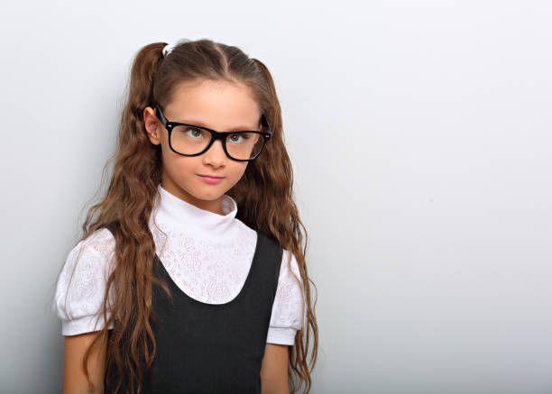 Grimacing  pupil girl with long hair style in fashion eyeglasses in uniform squint eyes on blue background with empty copy space. stock photo