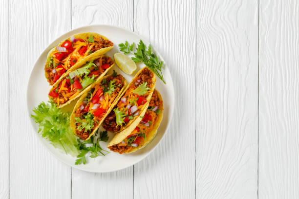 Ground Beef Tacos with shredded cheddar cheese, fresh lettuce, tomato, onion on a white plate with lime wedges, horizontal view from above, mexican cuisine, flat lay, free space stock photo
