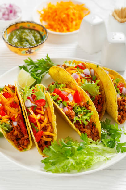 Ground Beef Tacos with shredded cheddar cheese, green salsa verde sauce, fresh lettuce, tomato, onion on a white plate with lime wedges, vertical view, close-up stock photo