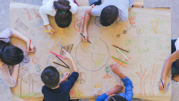 Pandangan overhead udara dari kelompok multi-etnis anak-anak usia dasar menggambar. Mereka duduk di sekitar meja. Anak-anak menggunakan pensil warna untuk membuat mural. Telah mewarnai peta dunia, benda-benda yang ditemukan di alam, dan simbol konservasi lingkungan.