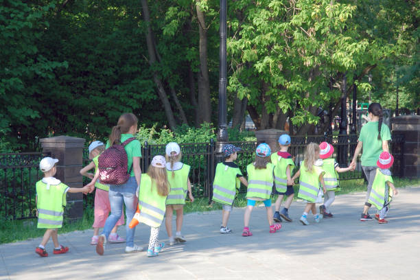 grupp av barn i hattar och mössor bär säkerhetsvästar går på trottoaren hand i hand med vuxna. dagis lärande trafiksäkerhetsregler utomhus på sommardagen - trafiklärare bildbanksfoton och bilder