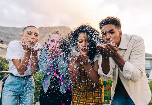 Group of friends blowing colourful confetti outdoors. Happy young people celebrating during a party on the rooftop. Multicultural friends having fun together on the weekend.