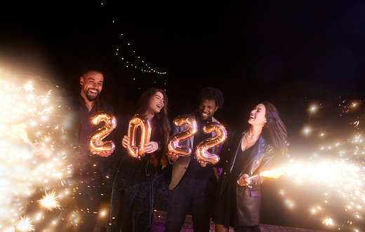 Group of multi-ethnic friends celebrating new years holding 2022 balloons and sparklers having fun at a outdoors party
