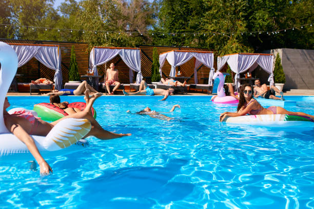 Photo of Group of friends chilling in private villa swimming pool lie in the sun on inflatable flamingo, swan, mattress. Young people relax with floaties at luxury resort on sunny day. Bikini girls sunbathing