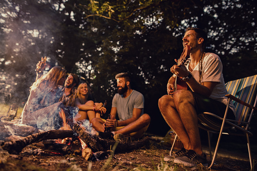 Young man sitting by the bonfire and playing acoustic guitar while his friends are having fun in the background.