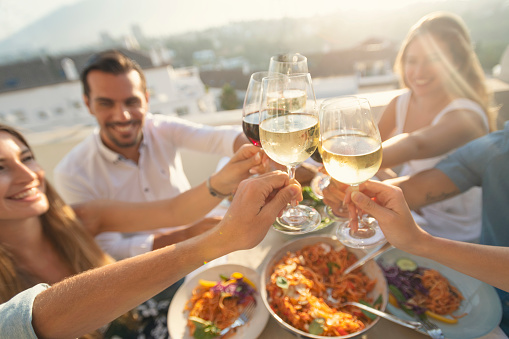 Group of friends having a meal outdoors. They are celebrating with a toast using wine. They are all happy and smiling. There are bowls of food on the table including salad and spaghetti Bolognese. Multi ethnic group.