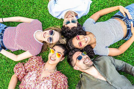 Group of friends lying on park meadow wearing colored trendy sunglasses looking at camera laughing. Happy diverse international people enjoying friendship in nature resting in circle. Mixed race union
