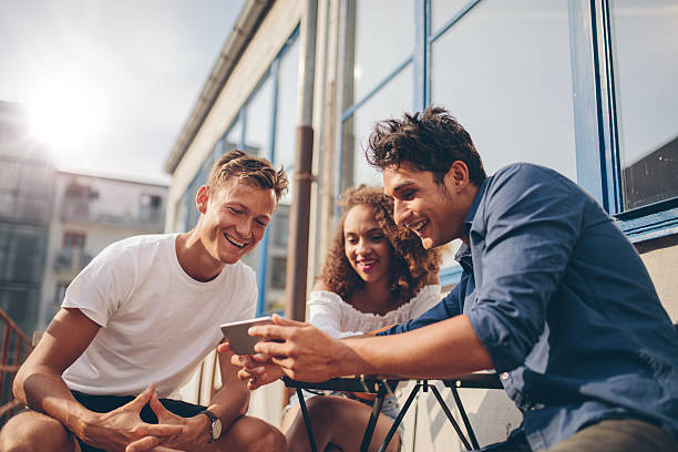Group of friends watching video on smartphone Three young friends sitting outdoors and looking at mobile phone. Group of people sitting at outdoor cafe and watching video on the smartphone. jacob ammentorp lund stock pictures, royalty-free photos & images