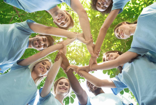Group of happy volunteers collaborate at park Support concept. Happy volunteers holding hands in circle and smiling, teambuilding exercise, teamwork and help, view from below, copy space dedication stock pictures, royalty-free photos & images