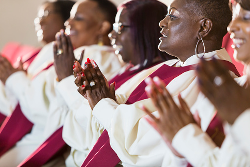A group of mature black women in church robes, sitting in a row, clapping. They are members of the church choir listening to a sermon.
