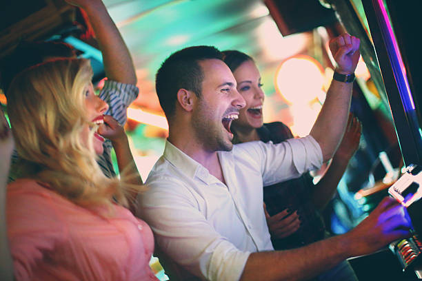Group of people playing slots. Group of young adults in mid 20's playing slots in a casino and having fun.Cheering,laughing...two couples on double date.Many slot machines stretch blurry in background. gambling stock pictures, royalty-free photos & images