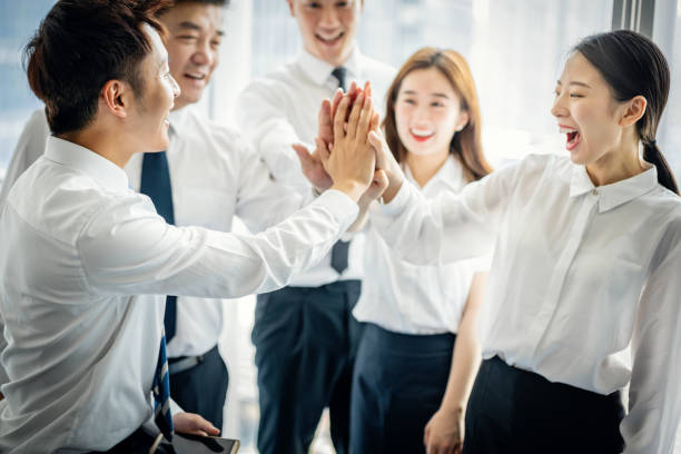 Group of smiling businesspeople high fiving each other while standing together in a modern office building stock photo