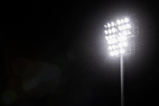 A group of glowing high intensity lamps to illuminate a sports field atop a steel pole at night with insects flying around against a black sky