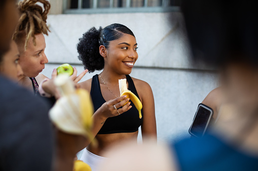Group of women eating banana in workout training break. Happy multi-ethnic females relaxing after completing exercise session standing outdoors and eating bananas in morning.