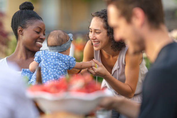 A group of young adult friends dining al fresco on a patio A young African American mom holds her adorable young daughter in her lap at an outdoor dining table. The girl is smiling and reaching toward mom's friend, who's smiling at her. barbecue meal photos stock pictures, royalty-free photos & images