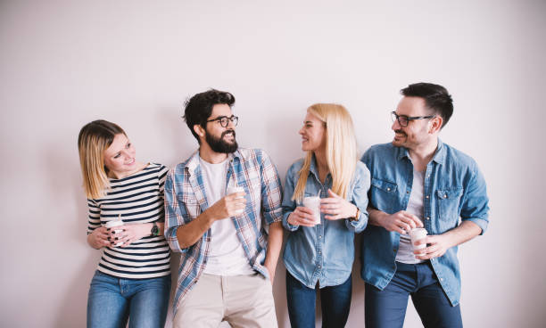Group of young stylish happy people leaning against the wall and talking while drinks coffee in the paper cup.  four people stock pictures, royalty-free photos & images