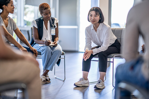 A small group of diverse adults sit together on chairs in a circle.  They are each dressed casually and are sharing their struggles with one another in a group therapy session.  A mature woman of African decent is leading the session as she allows a young Asian woman to share with the group.