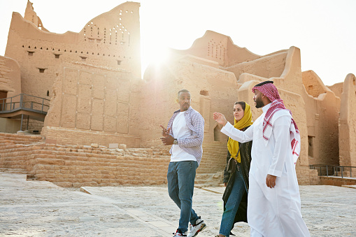 Three-quarter front view with lens flare of male and female tourists in their 30s listening to Saudi man in traditional attire describe history of At-Turaif. Property release attached.
