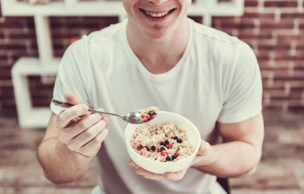 Guy with healthy food Handsome young guy is smiling while eating porridge with berries in kitchen at home dietary fiber stock pictures, royalty-free photos & images