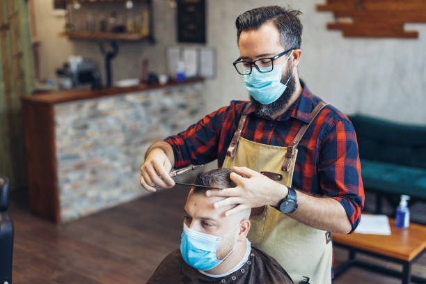 Hair cutting during pandemic. Young man have hair cutting at barber shop during pandemic isolation, they both wear protective equipment. stock photo