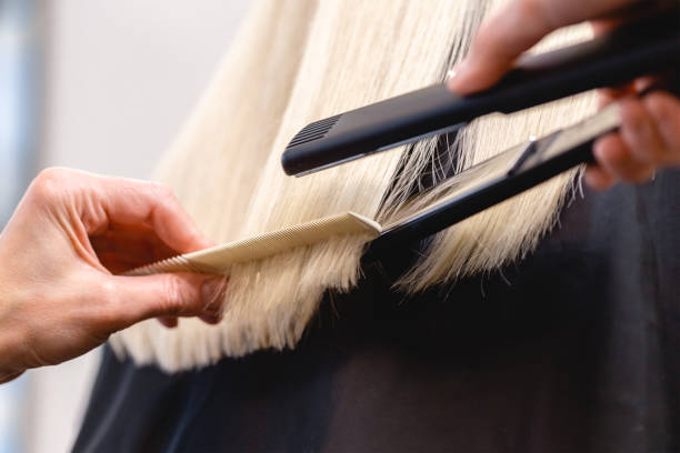 Hairdresser straightens blonde hair with flat iron stock photo
