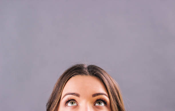 Half face of adorable woman's face looking up above copy space Closeup beautiful young woman looking up on blank copy space isolated on gray background. Attractive funny girl staring above her head. Half face of adorable woman thinking about solution. Empty space looking up stock pictures, royalty-free photos & images