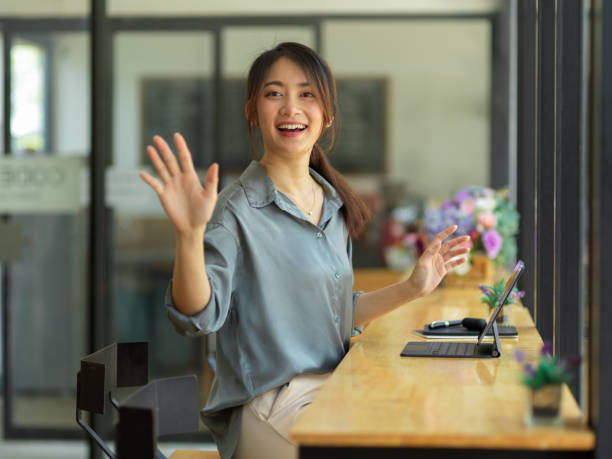 Half-length portrait of young beautiful female waving to say hi and smiling to camera Half-length portrait of young beautiful female waving to say hi and smiling to camera while sitting at workplace wave goodbye asian stock pictures, royalty-free photos & images