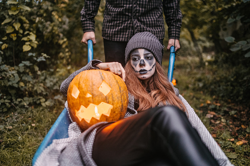Disguised woman with pumpkin and black balloon lying down in a construction trolley while man is pushing her in a yard. Halloween concept