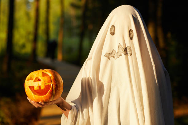Halloween Ghost Waist up portrait of funny little kid dressed as ghost holding pumpkin while posing outdoors on Halloween, copy space stage costume stock pictures, royalty-free photos & images
