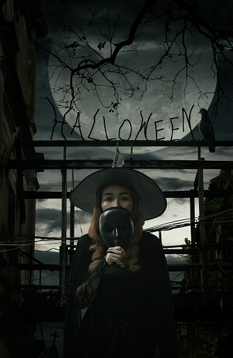 Halloween witch holding black face mask standing over damaged old wooden bridge, bird, dead tree, full moon with spooky cloudy sky, Halloween mystery concept